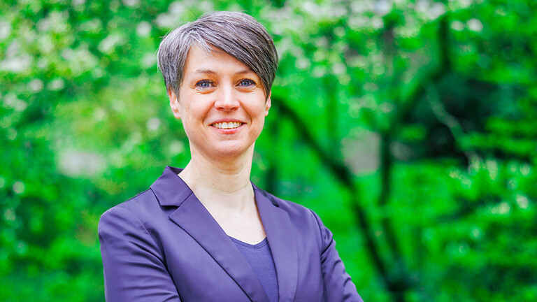 Portrait von Susanne Becker, sie hat kurze schwarze Haare, trägt ein lilafarbenes Jacket und steht draußen vor einer Baumreihe.