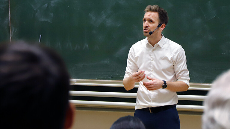 Julian Thimme, Professor am Karlsruhe Institute of Technology, hält eine Vorlesung bei der Nacht der Wissenschaft am Karlsruher Institut für Technologie. Er steht vor Publikum in einem Hörsaal. Im Hintergrund eine Tafel.