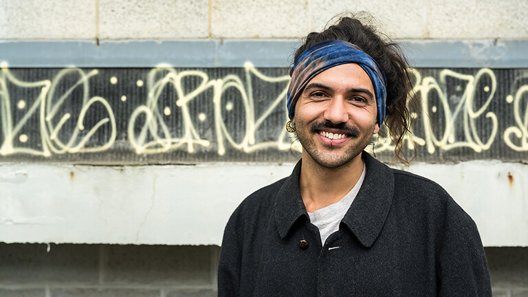 Portrait von Kevin Rozario. Kevin Rozario hat die langen Haare zusammengebunden. Trägt ein dunkles Hemd. Kevin Rozario blickt Richtung Kamera und lacht.
