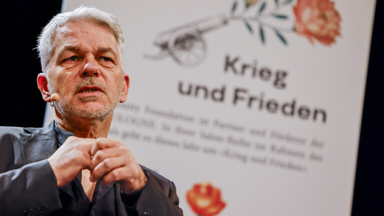 Carlo Masala auf einer Bühne, im Hintergrund ein Banner, auf dem "Krieg und Frieden" steht