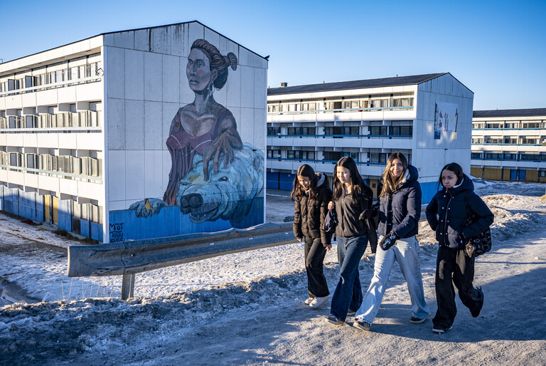 In der grönländischen Stadt Nuuk gehen Mädchen an Häusern vorbei. Es liegt Schnee. An einer der Hausfassaden ist ein großes Wandgemälde, das eine Inuk darstellt.