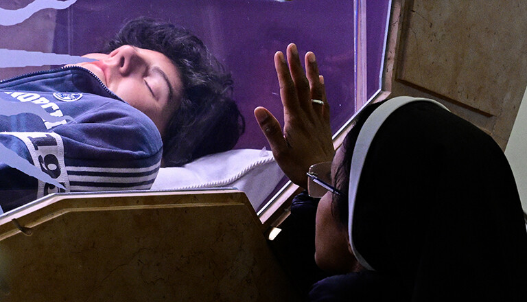 Foto einer Nonne, die ihre flache Hand auf die Seite des gläsernen Sarges von Carlo Acutis legt, der in der Kirche Santa Maria Maggiore zu Assisi steht.