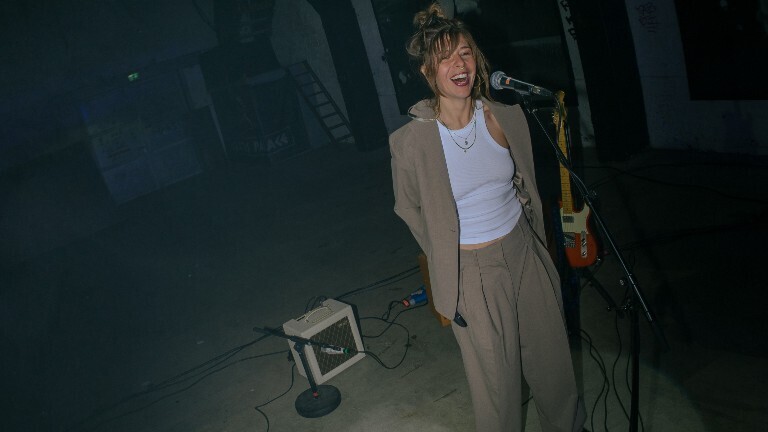 Musikerin Sophia steht vor einem Mikrofon und lacht dabei in die Kamera.