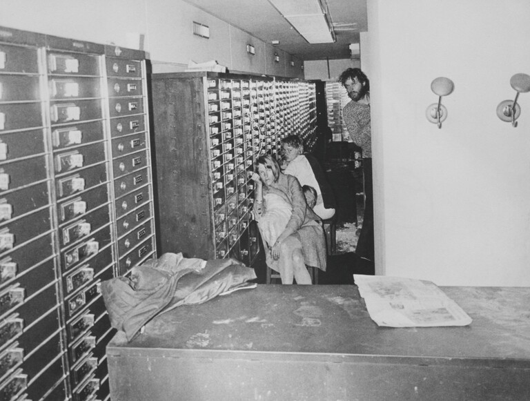 Archivfoto in schwarz-weiß: Blick in die Bank beim Überfall mit Geiselnahme im August 1973 in Stockholm. Zwei Menschen sitzen, rechts steht einer der Geiselnehmer.