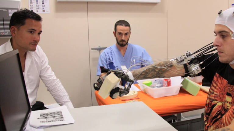 Foto des Neurotechnologen Surjo R. Soekadar (links) bei der Arbeit mit einem Patienten (rechts), der eine Elektrodenkappe trägt und am ausgestreckten Arm eine Exoskeletthand trägt, während er auf konzentriert auf einen Computerbildschirm guckt. Im Hintergrund beobachtet ein Mann in blauem Pflegerhemd (mittig) das Experiment.