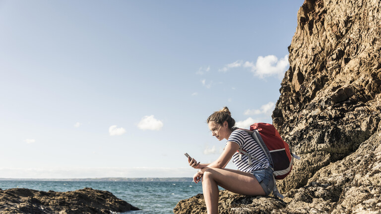 Junge Frau sitzt an einem felsigen Strand und benutzt ein Smartphone