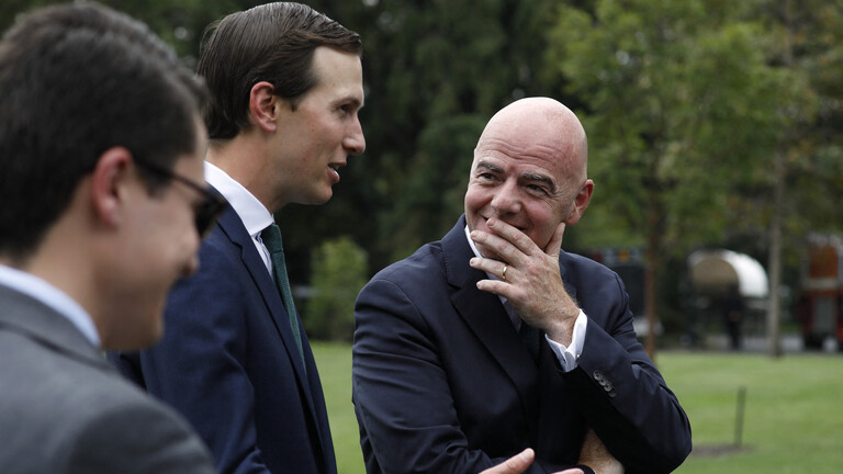 Zwei Männer in Anzügen unterhalten sich. Sie stehen auf einem Rasenstück. Eine unbekannte männliche Person links, über Jared Kushner und Gianni Infantino in der Mitte. Er schweigt und hat die linke Hand vor den Mund geführt. Eine Geste der Aufmerksamkeit.
