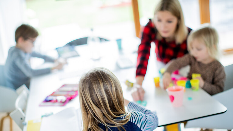 Eine Mutter sitzt an einem Tisch mit ihren drei Kindern. Sie machen Hausaufgaben oder spielen. Die Personen sind nur unscharf erkennbar.
