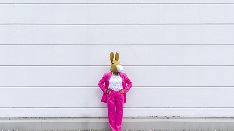 Eine Frau in einem pinken Anzug steht mit einer Eselsmaske vor einer weißen Wand