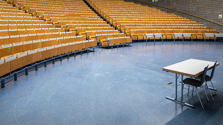 Blick in das leere Audimax der Technischen Universität Dortmund. Viele Stuhlreihen mit klappbaren Pulten links, rechts im Bild steht ein Tisch mit zwei Stühlen.