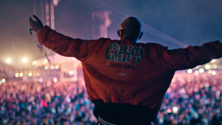 Filmstill der Netflix-Dokumentation "Babo – Die Haftbefehl-Story": Der Rapper Haftbefehl steht mit dem Rücken zur Kamera mit ausgebreiteten Armen auf einer Open-Air Bühne und schaut auf das große Publikum.