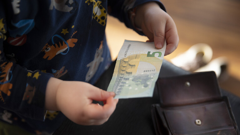 Ein Kind mit einem abgewetzten Geldbeutel hält einen 5-EUro-Schein in den Händen.