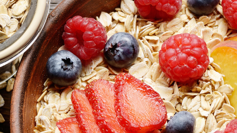 Draufsicht auf fruchtiges, gesundes Müsli mit Pfirsichen, Erdbeeren, Mandeln und Brombeeren in einer Tonschale auf einem Holzküchentisch.