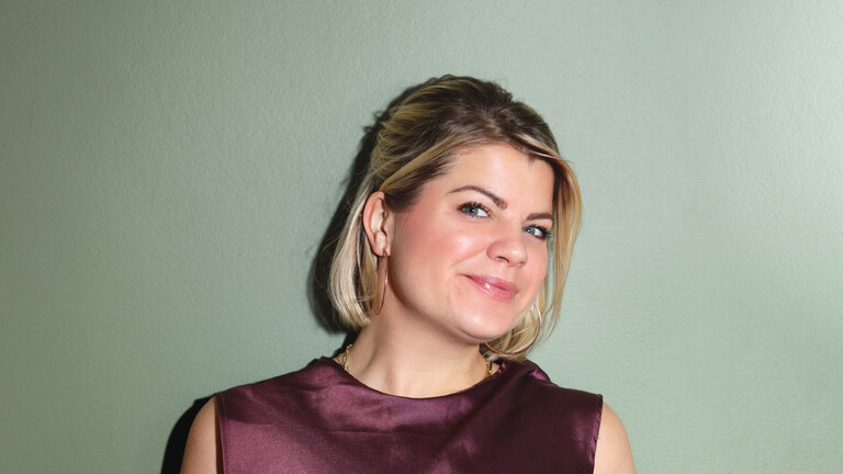 Frieda Lewin trägt ein dunkel bordeauxfarbenes Satin-Top und einen hellen engen Satinrock mit Pflanzenmotiven.