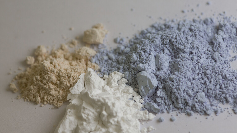 Auf einem Tisch liegen drei Pulverhaufen in braun, weiß und blau. Es sind die Seltenen Erden Ceroxid, Yttriumoxid und Neodymoxid.