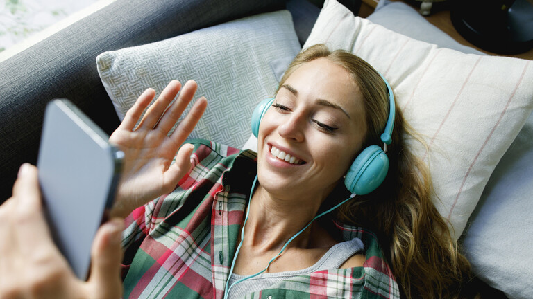 Frau liegt auf Sofa, sie trägt Kopfhörer und winkt lächelnd ins Smartphone