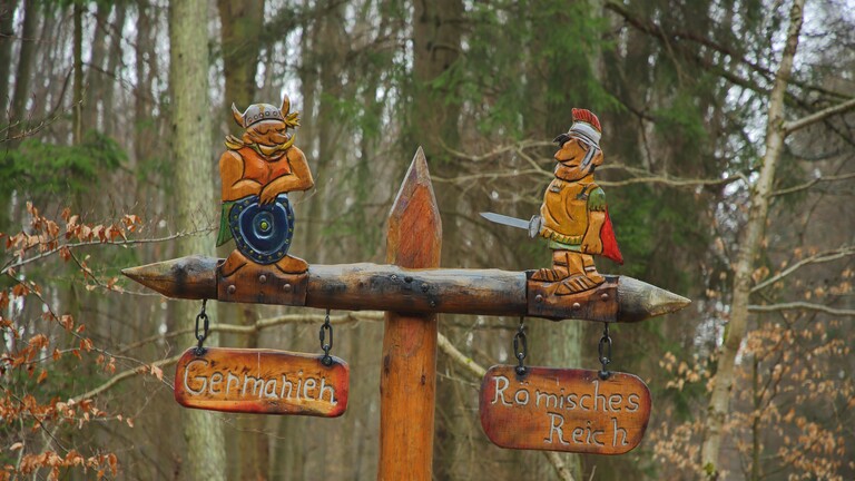 In Wetterau an historischen Limes-Grenze steht im Wald ein Hinweisschild: Oben drauf sind zwei Holzfiguren festgemacht, darunter hängen zwei kleine Holzschilder, darauf steht Germanien und Römisches Reich.