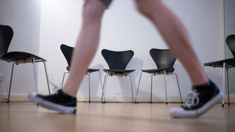 Sneaker tragende Person geht auf einem Gang an einer Reihe von Stühlen vorbei