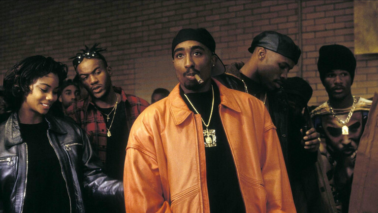 Der Rapper Tupac Shakur in einer Szene im Film Above The Rim aus dem Jahr 1994. Er trägt eine orangefarbene Bomberjacke und raucht eine Zigarre.
