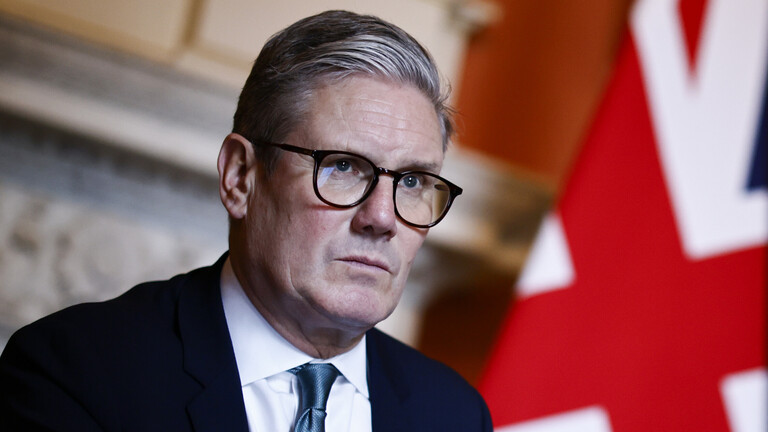 Der britische Premierminister Keir Starmer sitzt vor einer britischen Fahne. Er trägt Anzug und Schlips. Er blickt in die Ferne.