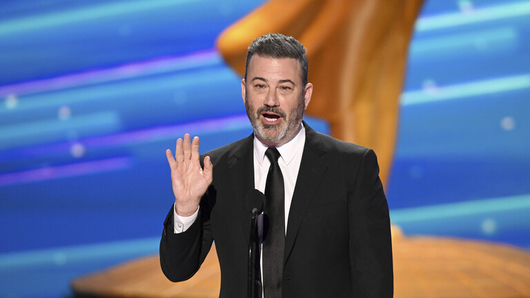 Ein Mann im schwarzen Anzug steht auf einer Bühne vor einem Mikrofon. Er hebt seine rechte Hand. Im Hintergrund ist viel Gold und Blau.