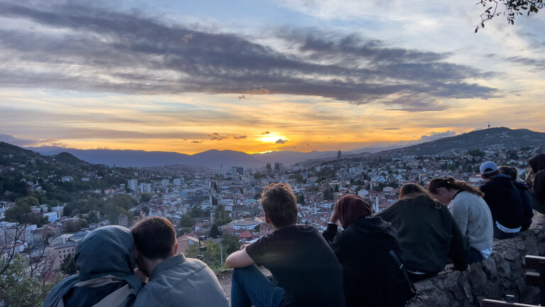 Touristen und Besucher blicken von einer Anhöhe auf und über die Stadt Sarajevo, die Sonne geht unter.