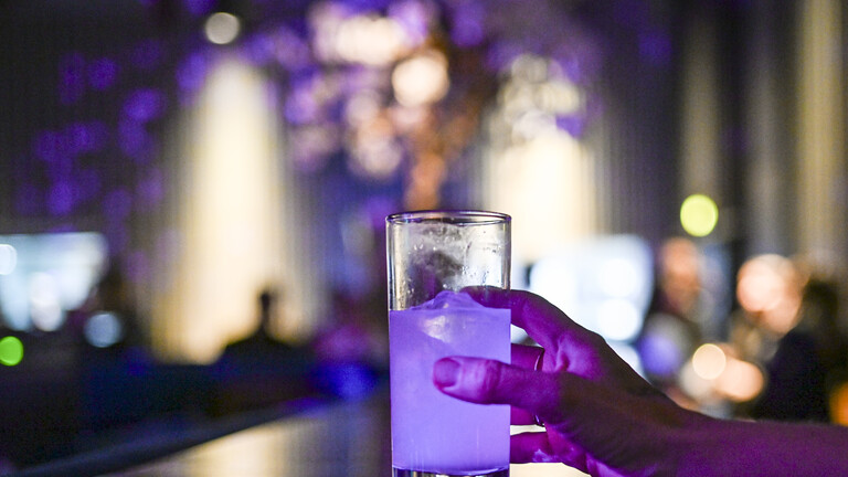 Die Hand einer Frau greift nach einem Glas mit einem Cocktail. Das Glas steht auf einer Theke, im Hintergrund ist unscharf eine Bar oder ein Club erkennbar.