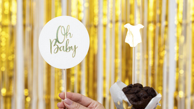 Eine Hand hält ein Schild mit der Aufschrift "Oh Baby", die andere Hand hält einen Schokomuffin in dem ein Babybody aus Papier steckt