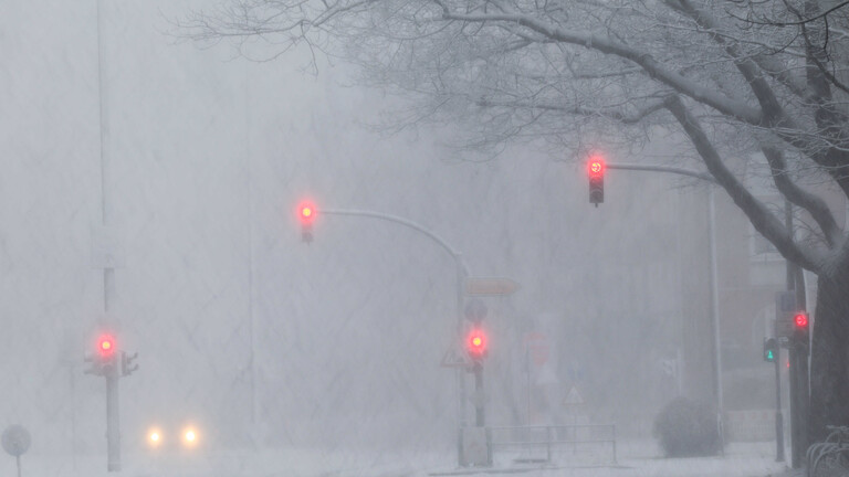 Scheinwerfer eines Autos und rot leuchtende Ampeln sind auf einer schneebedeckten Verkehrsstraße