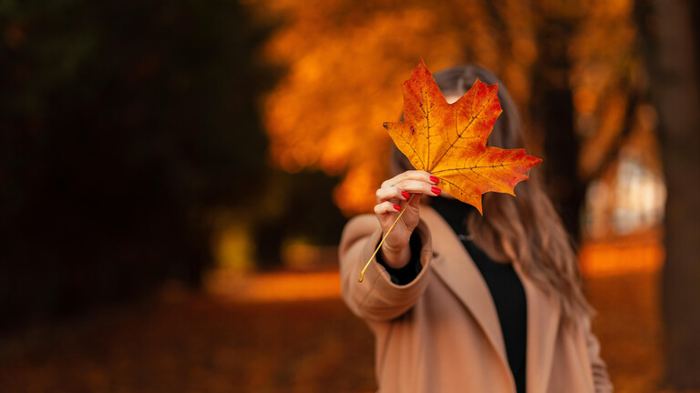 Frau in einem beigen Mantel steht im Park, wo sie sich ein großes, gelbes Herbstblatt vors Gesicht hält