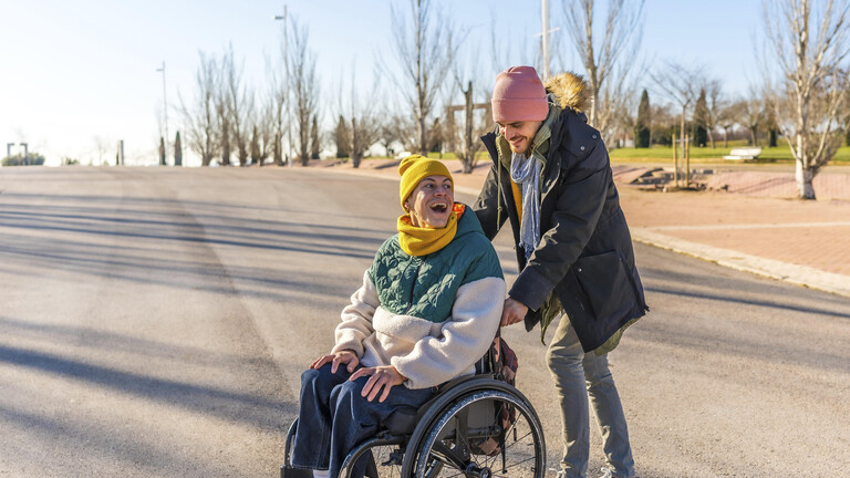 Junger Pfleger hilft einer fröhlichen älteren Frau im Rollstuhl, die einen sonnigen Wintertag in einem Park genießt
