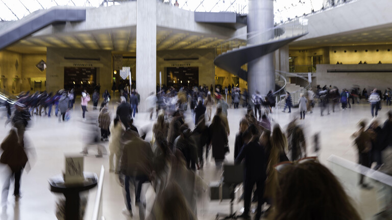 Viele Menschen stehen in einem hohen Raum, eine Wendeltreppe führt nach oben. Ihre Bewegungen sind unscharf. Es ist die Glaspyramide, Haupteingang zum Museum Louvre.