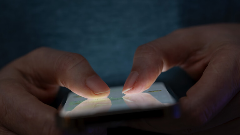 Das Bild zeigt eine Person, die in einer schlecht beleuchteten Umgebung ein Smartphone benutzt. Die Beleuchtung hebt die Finger des Benutzers und den Bildschirm des Smartphones hervor.
