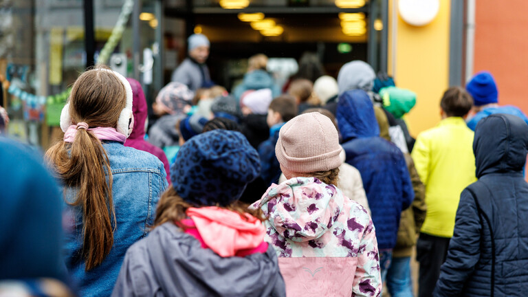 Schüler und Schülerinnen einer Grundschule gehen vom Schulhof ins Gebäude. Sie sind von hinten zu sehen. Sie tragen Jacken und teils Mützen.