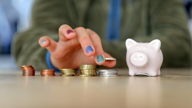 Hand mit lackierten Fingernägeln stapelt Geldmünzen neben einem Sparschwein