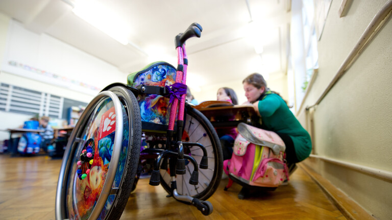 Ein Rollstuhl steht im Klassenzimmer, dahinter sitzen zwei Schülerinnen an einem Tisch