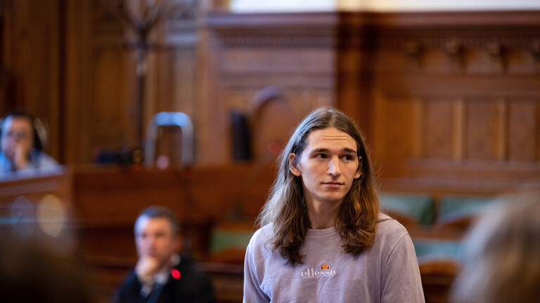 Maja T. steht im Gerichtssaal des Stadtgericht in Budapest. Die Wände sind holzvertafelt. Maja T. steht, Maja T. hat braune, lange Haare und trägt einen fliederfarbenen Pullover.
