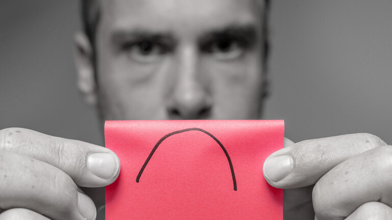 Ein Mann in Schwarz-Weiß hält einen roten Zettel in die Kamera, auf dem ein symbolischer Mund mit heruntergezogenen Mundwinkeln aufgemalt ist.