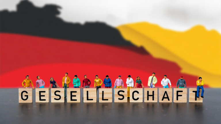 Fotomontage, Menschen stehen hinter Holzbuchstaben mit dem Wort „Gesellschaft“ vor einer Deutschland-Flagge /  Sinnbild für nationale Identität, Vielfalt, Zusammenhalt und die Bedeutung gemeinsamer Werte in Deutschland.