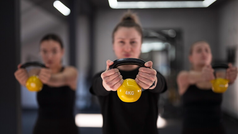 Gruppe von Frauen führen Kesselbell Übungen in einem gut beleuchteten Fitness-Studio. Sie konzentrieren sich auf ihre Form, während sie gelbe Kesselbells halten und passende Outfits tragen.