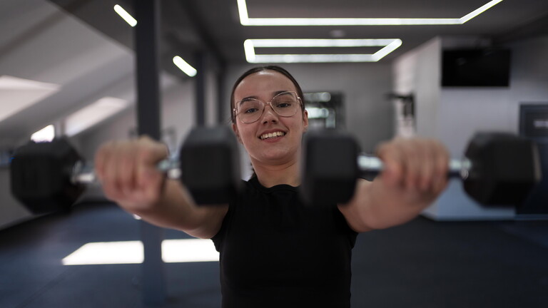 Eine junge Frau steht im Fitness-Studio. Sie blickt in die Kamera, sie hebt zwei Hanteln in die Höhe.
