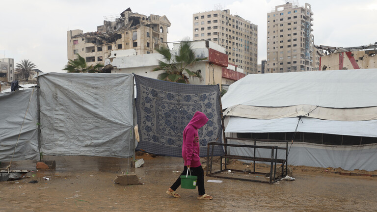 Ein Palästinenser trägt einen Eimer voll mit Trinkwasser in einer von den Überschwemmungen betroffenen Notunterkunft aus Zelten. Im Hintergrund sind zerstörte Häuser zu sehen.