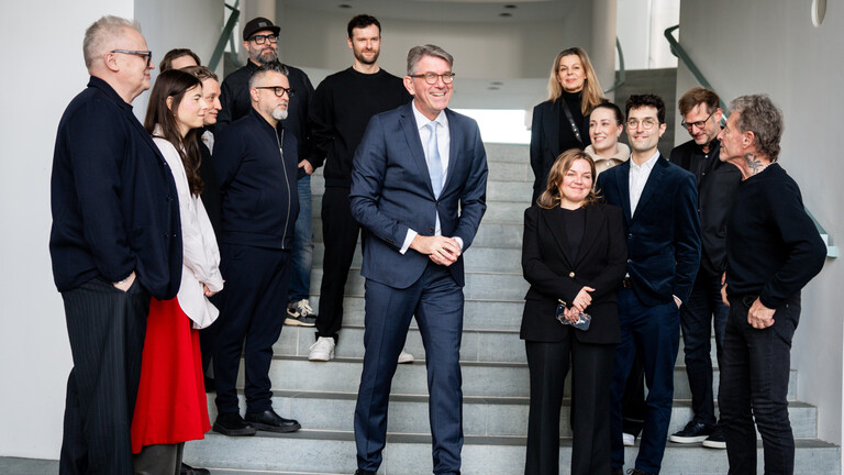 Kulturstaatsminister Wolfram Weimer steht im Bundeskanzleramt vor einer Treppe. Um ihn und auf der Treppe stehen verschieden Musiker, unter anderem Herbert Grönemeyer.
