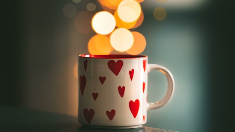 Auf einer weißen Tasse sind rote Herzen abgebildet. Die Tasse steht auf einem Tisch, im Hintergrund sind gelbe Lichtreflexe erkennbar.