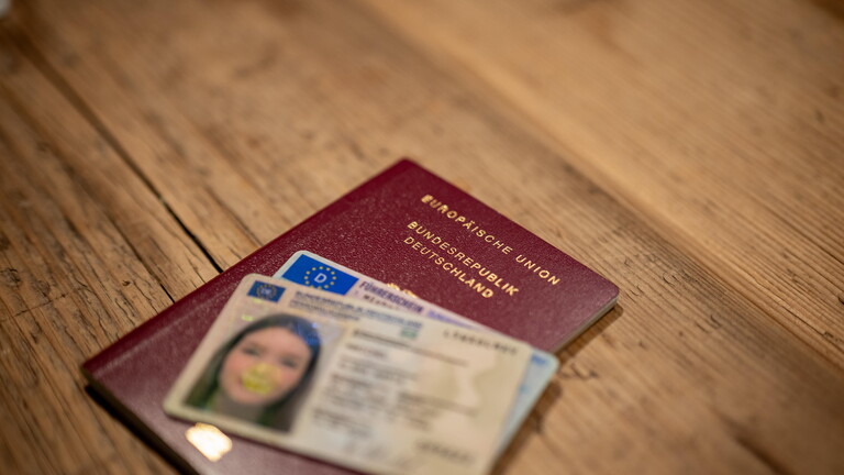 Eine Hand hält einen deutschen Reisepass zusammen mit einem Führerschein über einem Holztisch.