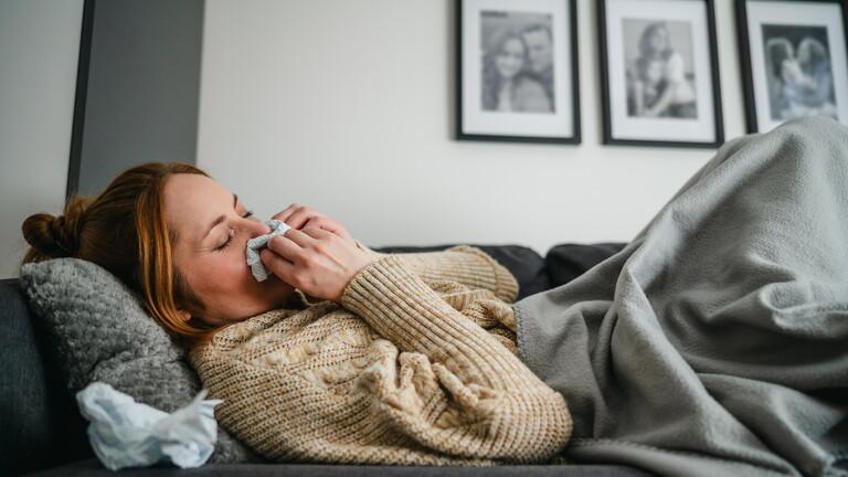 Frau liegt mit Decke auf Sofa und putzt sich die Nase