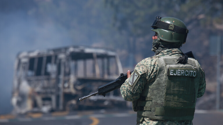 Ein Soldat mit Maschinengewehr steht vor einem ausgebrannten Fahrzeug auf einer Straße in Cointzio, Mexiko (22.02.2026)