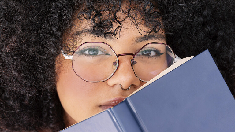 Eine Frau mit lockigem Haar schaut über ein blaues Buch und trägt eine große runde Brille. Ihr Gesichtsausdruck ist konzentriert und unterstreicht ein Gefühl von Neugier und Kontemplation