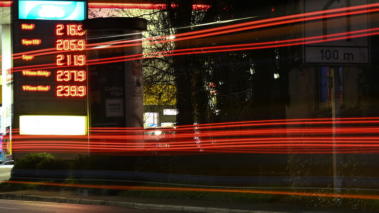 Die Scheinwerfer vorbeifahrender Fahrzeuge ziehen am Morgen auf einer Straße Lichtspuren, während im Hintergrund die Preistafel einer Tankstelle die Kraftstoffpreise anzeigt.