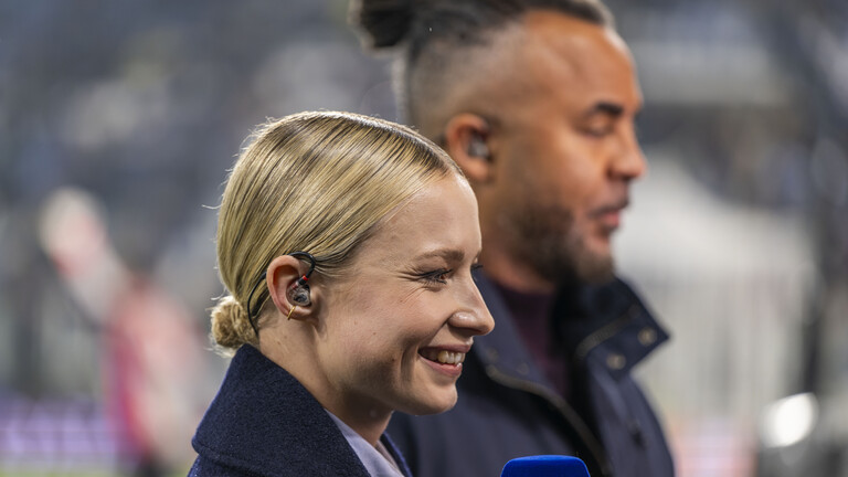 Sportmoderatorin Lena Cassel hält ein Mikrofon in der Hand. Sie steht mit Experte Patrick Owomoyela bei einem Championsleague-Spiel auf dem Fußballplatz.